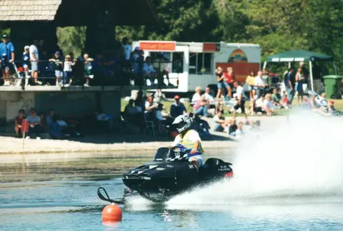 A person rides a snowmobile across a body of water in front of spectators.