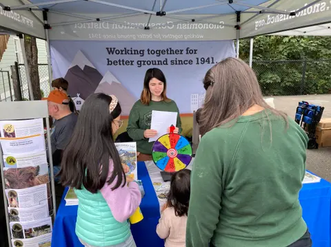 A woman stands behind a booth at an outdoor event, talking to people.
