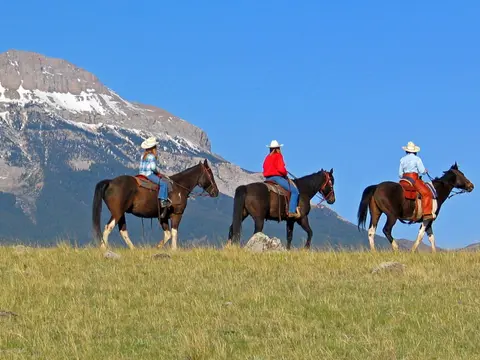 Three people ride horses across a grassy field in front of a mountain.