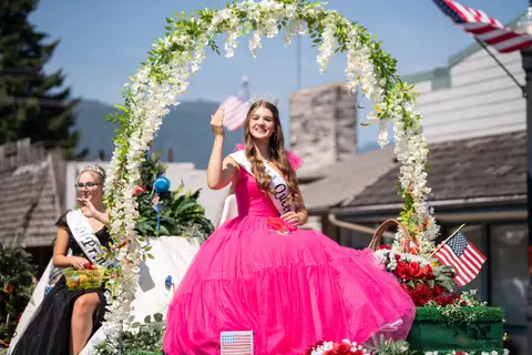 A young woman wearing a pink dress and tiara waves from a parade float decorated with flowers and American flags.