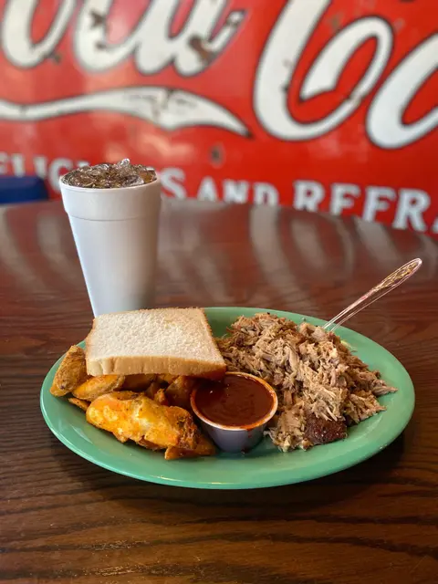 A plate of pulled pork, potato wedges, bread, and sauce next to a drink on a wooden table.
