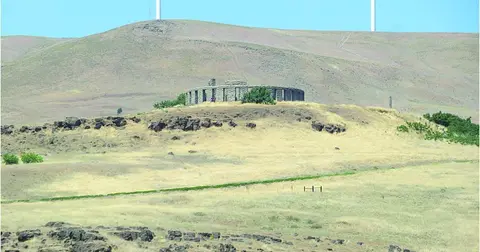 A stone structure sits on a grassy hill with hills and wind turbines in the background.