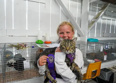 A young person holds a cat and a ribbon inside a building with cages.