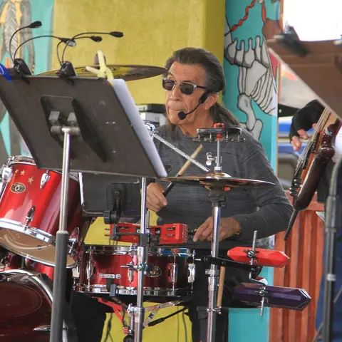 A drummer plays a drum kit outdoors with a colorful mural in the background.