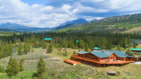 An aerial view of a wooden lodge nestled in a scenic valley with mountains in the background.