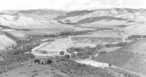 Black and white landscape photograph showing a valley with mountains in the background and figures with horses in the foreground.