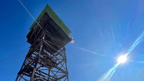 A tall wooden fire lookout tower stands under a bright sun and blue sky.