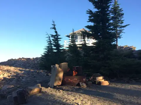 A campfire burns in a metal ring with a small building on a rocky ridge behind it.
