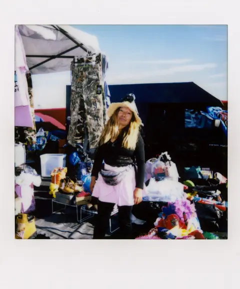 A woman stands in front of a market stall displaying clothes and items.