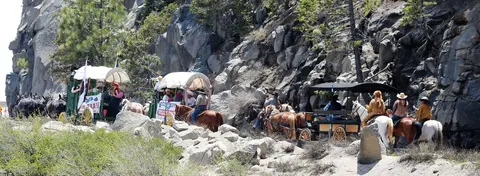 A wagon train with horses and carriages travels along a rocky mountain road.