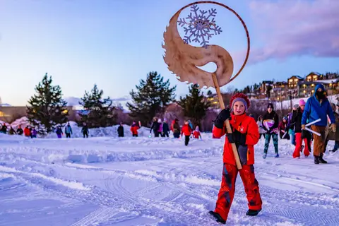 A child in winter gear carries a decorative wooden symbol during a snowy outdoor event.