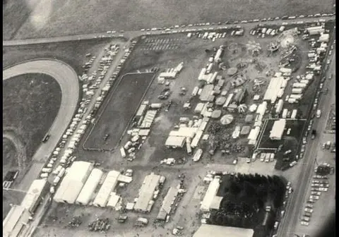 An aerial black and white photograph showing a fairgrounds with carnival rides, exhibition buildings, and parking lots.
