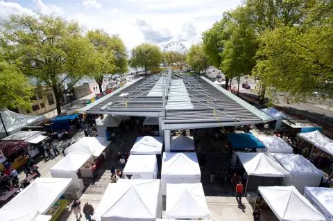 An elevated, high-angle outdoor shot shows a large open-air market with many white tents and people walking underneath a modern canopy roof near a waterfront, with a Ferris wheel in the distance.