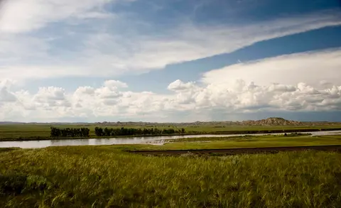 A river flows through a grassy plain under a partly cloudy sky.