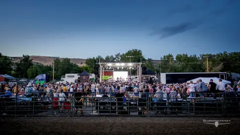 An outdoor concert performance viewed from behind a metal barrier with a large crowd attending.