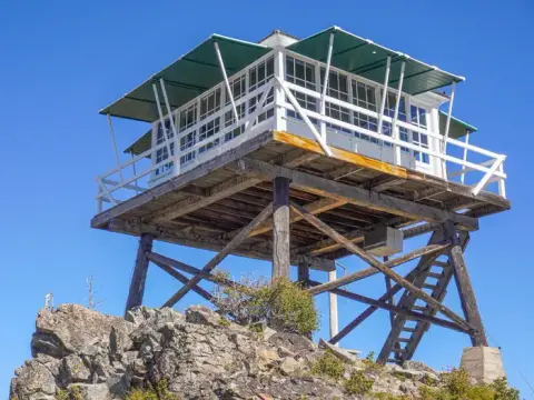 A fire lookout tower stands atop a rocky peak against a clear blue sky.