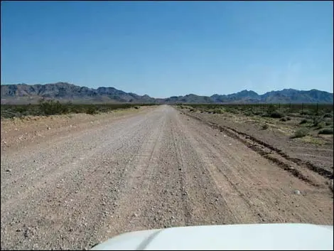 A view of a dirt road stretching through a desert landscape toward distant mountains.