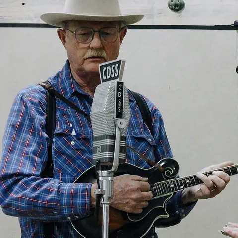 A man wearing a cowboy hat and plaid shirt plays a mandolin in front of a vintage microphone.
