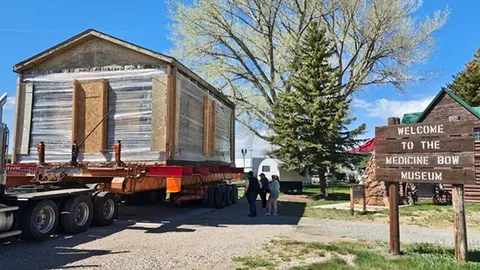 A large building being transported on a flatbed trailer near a wooden sign and trees.