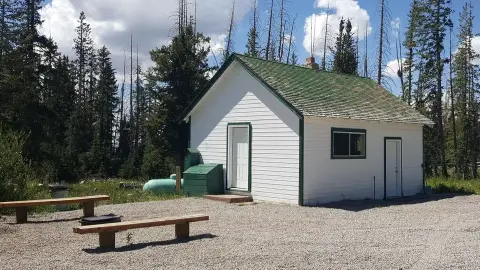 A small white building with a green roof stands in a clearing surrounded by trees.