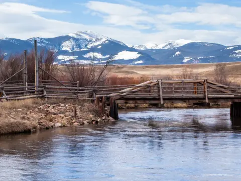 A wooden bridge crosses a river with snow-capped mountains in the background.