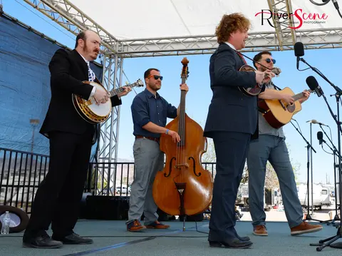 A four-piece band performs on an outdoor stage under a white canopy.