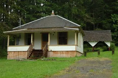 A small white house with a porch and a detached covered structure set among trees.