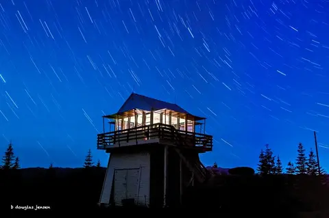 A fire lookout tower stands under a blue night sky with star trails.