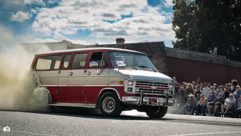 A vintage van performs a burnout on a street as a crowd watches.