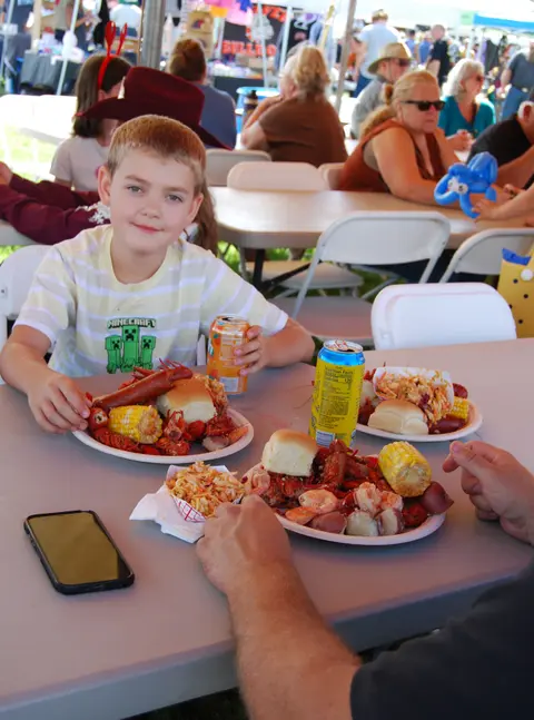 A young boy sits at an outdoor table with a plate of seafood and other food items.