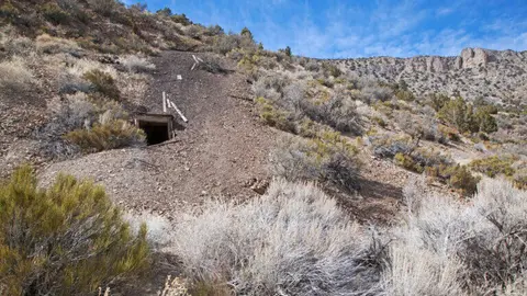 A mine shaft opening built into a hillside covered with brush and rocks under a blue sky.