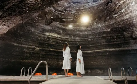 Two women in white robes stand on a wooden dock inside a rocky cave with a pool.