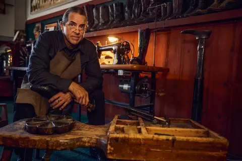 A craftsman poses in a workshop filled with boots and shoemaking tools.