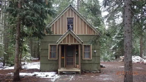 A small cabin stands surrounded by tall trees in a snowy forest.