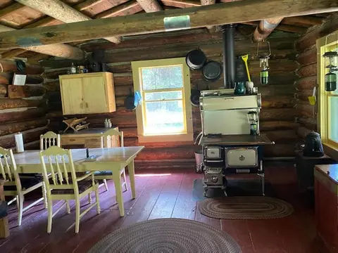 An interior view of a rustic log cabin with a wood-burning stove and dining table.