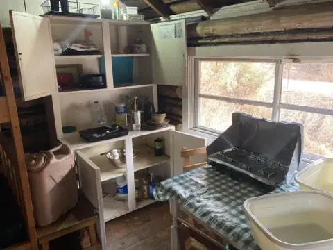 An interior view of a rustic cabin kitchen with open cupboards, a table, and a camp stove.