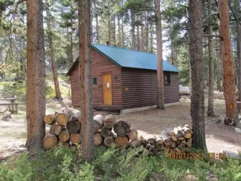 A wooden cabin surrounded by trees with stacked logs in the foreground.