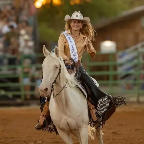 A woman wearing a cowboy hat and sash rides a white horse in an arena.