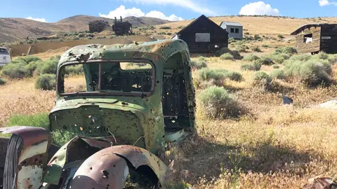 An old ruined vehicle sits in a field with wooden buildings in the background.