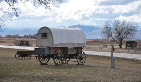 A covered wagon parked on a grassy field with mountains in the background.