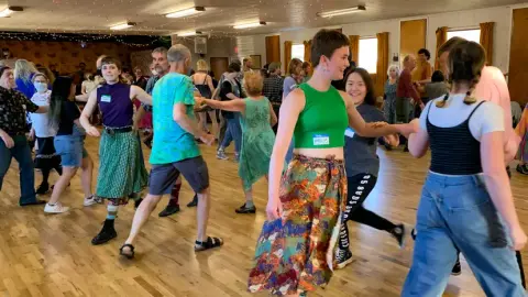 A group of people dancing together inside a hall with wooden floors.