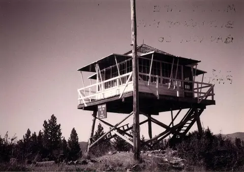 Historic black and white photograph of a fire lookout tower on a wooden stilt structure.