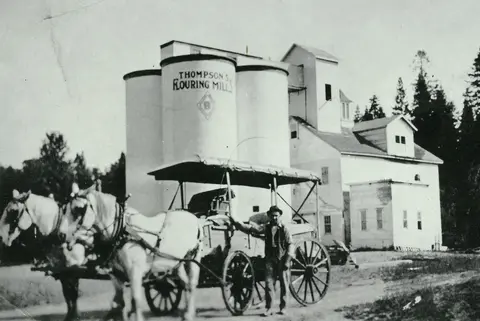 Historic photo of a horse-drawn wagon outside a flouring mill with silos.