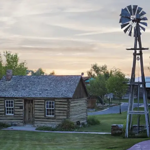 A rustic log cabin and a tall metal windmill stand in a grassy area at sunset.