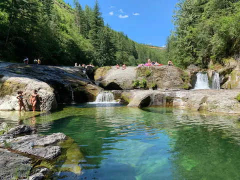 People swim and relax in a clear natural pool surrounded by rocks, waterfalls, and forests.