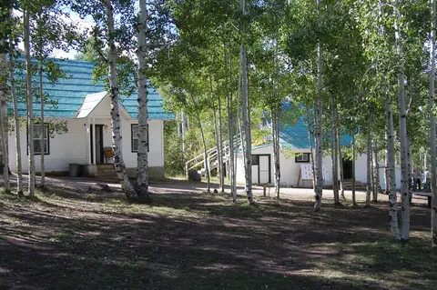 White buildings with teal roofs stand among a grove of aspen trees.
