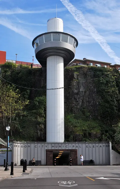 A tall modern municipal elevator stands against a rock cliff with people nearby.