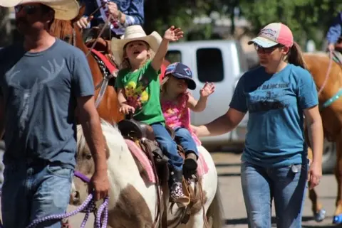 Two children ride a pony led by adults at an outdoor event.