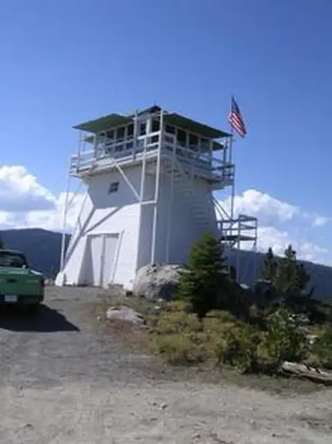A white fire lookout tower sits on a mountaintop with an American flag flying.