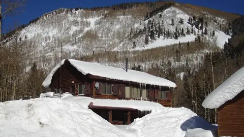 A snow-covered log cabin sits in front of a large snow-covered mountain.